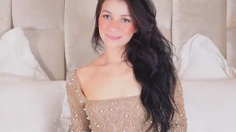 Media: Video of a young woman with long, wavy black hair, fair skin, and light makeup, wearing a gold sequined off-the-shoulder dress, seated on a white bed with a beige headboard in a softly lit bedroom.