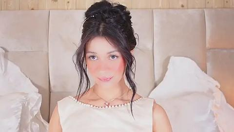 Media: Video of a young woman with light skin, dark hair in a bun, and rosy cheeks, wearing a white sleeveless dress, standing against a beige tufted headboard with white pillows.