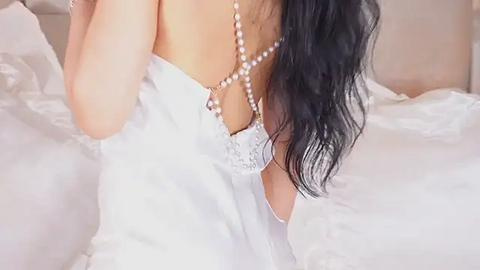 Media: Video of a woman with long, wavy black hair, wearing a white lace bra and pearl necklace, partially covered by a white sheet, lying on a bed with white linens.