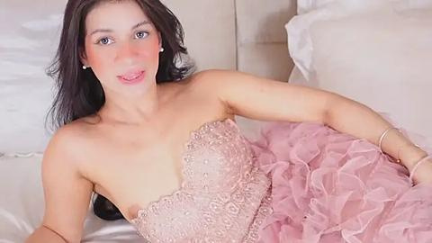 Media: Video of a fair-skinned woman with long black hair, wearing a pink ruffled gown, lying on white bedding, with a relaxed expression.