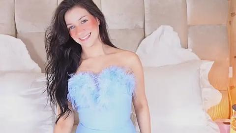 Media: Video of a smiling, light-skinned woman with long black hair, wearing a strapless, blue dress with fluffy sleeves, standing in a bedroom with white bedding and a beige headboard.