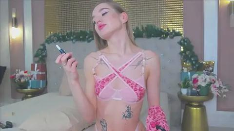 Media: Video of a slender, fair-skinned, blonde woman with tattoos, wearing pink lingerie with crisscross straps, holding a bottle, in a modern, festive bedroom with floral decorations.