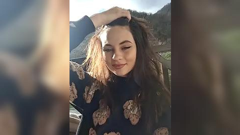 Media: Video of a young woman with long, wavy dark hair, wearing a black, patterned sweater, smiling and running her hand through her hair, with a blurred mountainous background.