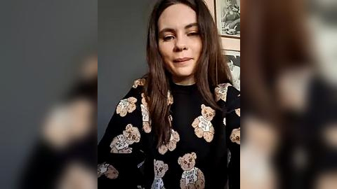 Media: Video of a young woman with long brown hair, wearing a black sweater adorned with teddy bear patterns, standing indoors. Background features a blurred, textured wall with framed art.