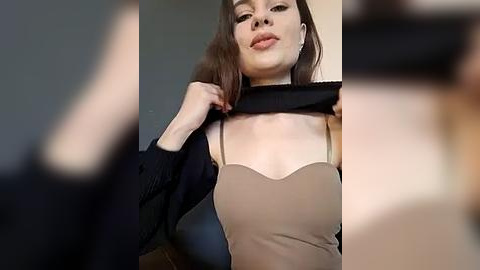 Media: Video of a light-skinned woman with straight brown hair, wearing a black sweater and beige top, pulling at her neckline, set against a blurred background.