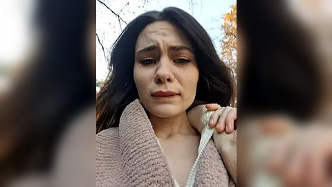 Media: Video of a woman with long, dark hair, wearing a pink, textured robe, looking distressed, holding a white cord, blurred background with autumn trees.