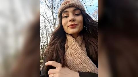 Media: Video of a young woman with long dark hair, wearing a beige knit hat and scarf, smiling outdoors amidst leafless trees on a cloudy day.