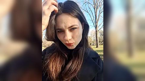 Media: Video of a young woman with long brown hair, wearing a black coat, making a pouty face outdoors in a park with bare trees and blue sky.