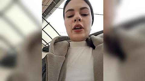 Media: A close-up video of a Latina woman with light brown skin and dark hair, wearing a beige coat, looking down thoughtfully. Background shows a blurred, beige-tiled room.