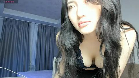 Media: Video of a young woman with long, wavy black hair, fair skin, and full lips, wearing a black lace bra, leaning forward, in a dimly lit bedroom with grey curtains.