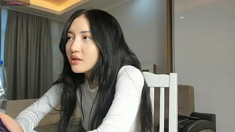 Media: Video of an East Asian woman with long black hair, fair skin, and a slim build, wearing a light grey sweater, sitting indoors, looking thoughtful.