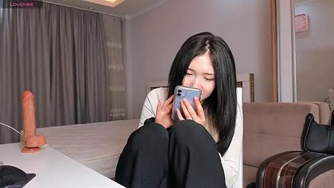 Media: Video of an East Asian woman with straight black hair, wearing a white top and black pants, sitting on a white couch, holding a phone, in a modern living room with beige curtains, a large dildo, and a brown blanket.