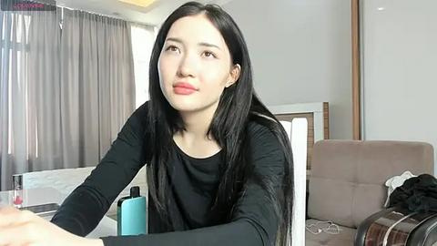 Media: Video of a young East Asian woman with long black hair, fair skin, and red lipstick, sitting in a modern living room with beige furniture and gray curtains.