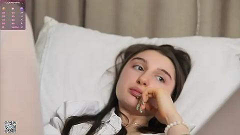 Media: Video of a young Caucasian woman with long dark hair, wearing a white blouse, lying on a bed, looking thoughtful with a hand to her mouth.