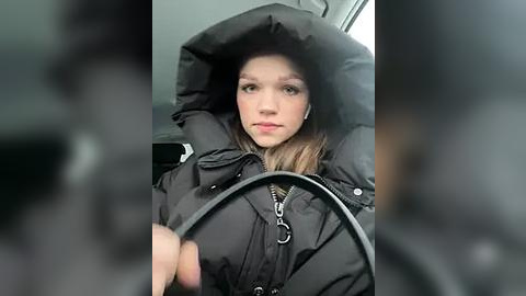 Media: Video of a young woman with fair skin, brown hair, and light makeup, wearing a black puffy winter coat and headphones, driving in a car.