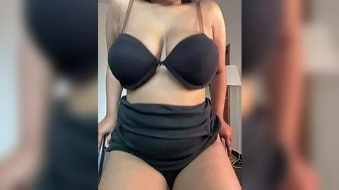 Media: Video of a curvy, light-skinned woman in a black bra and high-waisted shorts, with her hands on her hips, indoors, blurred background.
