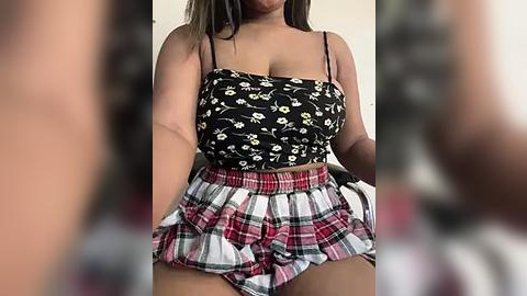 Media: Video of a woman with medium skin tone, wearing a black floral crop top and a short, plaid skirt, standing indoors with blurred background.