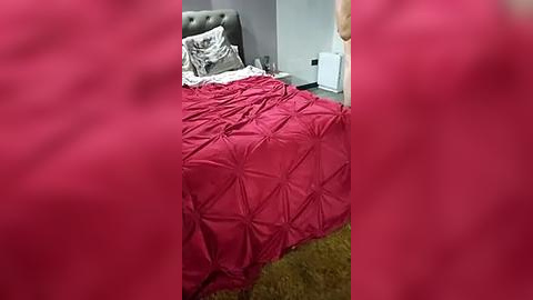 Media: Video of a tidy bedroom with a tufted red bedspread, gray upholstered headboard, white pillows, beige carpet, and a white nightstand beside a closed door.