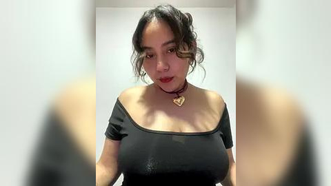 Media: Video of a young Asian woman with wavy black hair, wearing a black off-shoulder top, a red choker, and a heart pendant, standing indoors with blurred background.