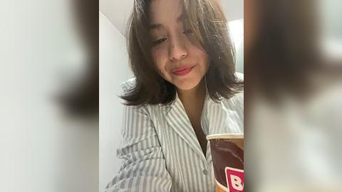 Media: Video of a young woman with shoulder-length dark hair, wearing a white and blue striped button-down shirt, holding a Dunkin' Donuts coffee cup. She has a neutral expression.