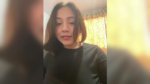 Media: A video of a young Asian woman with shoulder-length dark hair, wearing a black shirt, standing in a brightly lit room with a floral-patterned curtain.