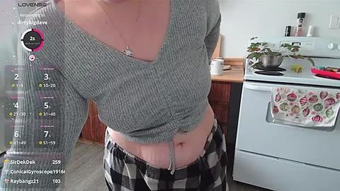 Media: A video of a woman with fair skin wearing a grey ribbed crop top and black-and-white plaid pajama pants, standing in a modern kitchen with a stove, sink, and potted plant.