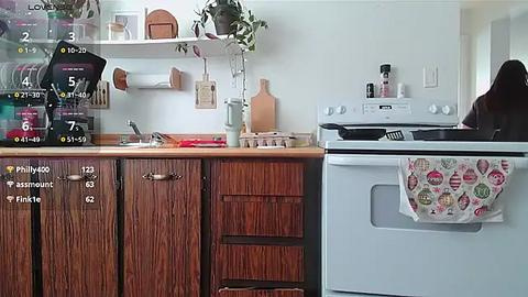 Media: Video of a cozy, rustic kitchen with wooden cabinets, a black coffee maker, a stove, and a hanging pot rack, showcasing a woman in a white shirt.