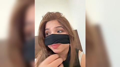 Media: A video shows a young woman with light brown hair, wearing a black mask and a white top, sitting with a playful expression, her hand near her mouth. The background is blurred.