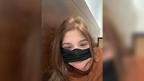 Media: Video of a young woman with light skin, long brown hair, wearing a black face mask and a brown sweater, standing in a dimly lit room with a wooden door and white ceiling.