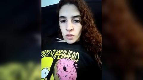 Media: Video of a young woman with curly brown hair, wearing a black \"South Park\" t-shirt, seated in a dark car. Her expression is neutral. Background is blurry.