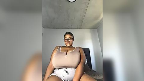 Media: Video of a plus-sized Black woman with dark skin, glasses, and a beige tank top, sitting on a chair in a minimalistic room with a concrete ceiling and a grey wall.