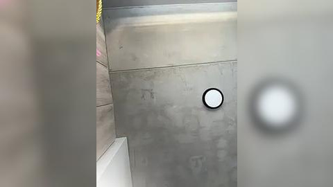 Media: Video of a modern bathroom with a circular light fixture on a grey concrete wall, featuring a yellow rope and a blurry, partially visible person in the background.