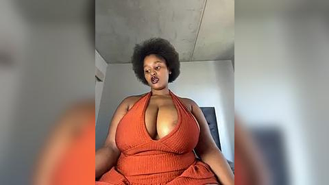 Media: Video of a plus-sized Black woman with large breasts, wearing a revealing orange halter dress, sitting in a modern room with concrete ceiling and white walls.