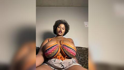 Media: Video of a plus-sized Black woman with natural hair, wearing a colorful, revealing top and shorts, seated on a bed in a dimly lit room.