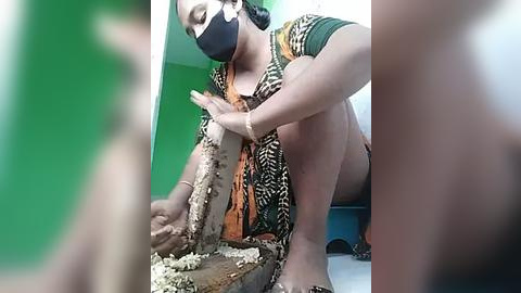 Media: Video of a dark-skinned woman wearing a mask, green shirt, and orange headscarf, squatting to clean a large, dirty snake on a green wall.