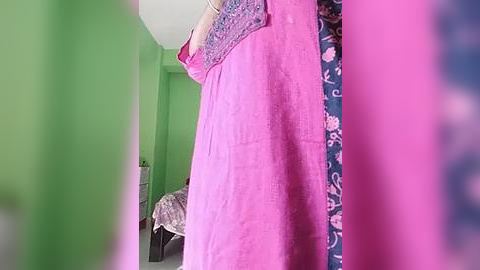 Media: A video of a bedroom with a bright pink dress hanging on a wall, green walls, a bed with floral bedding, and a blue-patterned shirt in the background.