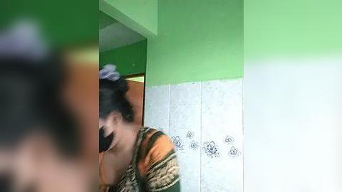 Media: A video of a person in a green and white bathroom with a blurry, partially obscured face. They wear a black face mask, and their hair is tied up. The background features a green wall and white tiled lower section.