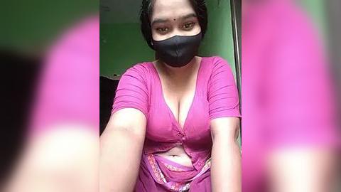 Media: Video of a young South Asian woman with medium brown skin, dark hair, and a black mask, wearing a pink top and colorful skirt, captured in a green-walled room with blurry foreground.