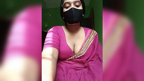 Media: Video of a South Asian woman in a pink sari with a black mask, holding out her arms. Background shows a blurred green wall and doorway.