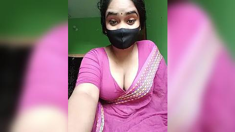 Media: Video of a South Asian woman with medium skin tone, wearing a pink sari with a low-cut V-neck, black face mask, and a bindi. Background features green walls and a black cushion.