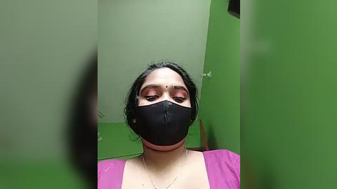 Media: Video of a South Asian woman with short black hair, wearing a black face mask, pink blouse, and green walls.