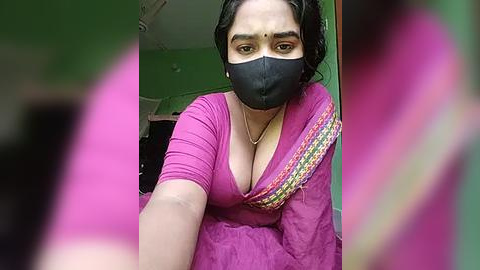 Media: A video of a woman with medium skin tone, wearing a black face mask, a pink blouse, and a colorful saree, with blurred figures in the background.