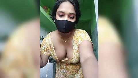 Media: Video of a South Asian woman with medium brown skin, short black hair, and a black face mask. She wears a low-cut floral top, revealing ample cleavage, and has a gold necklace. The background features a green wall and a shelf.