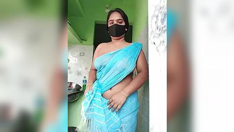 Media: Video of a plus-sized, dark-skinned woman with long black hair in a light blue sari, wearing a black face mask, standing in a green-walled room with a sink, a mirror, and white tiles.