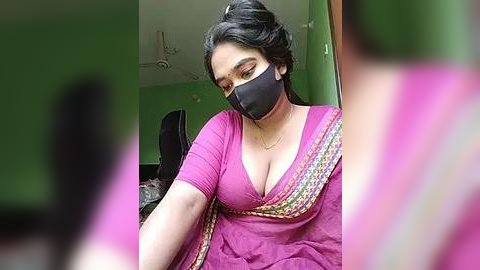 Media: Video of a South Asian woman with medium brown skin, wearing a black face mask, pink blouse, and colorful sari, standing in a green-painted room, blurred background.