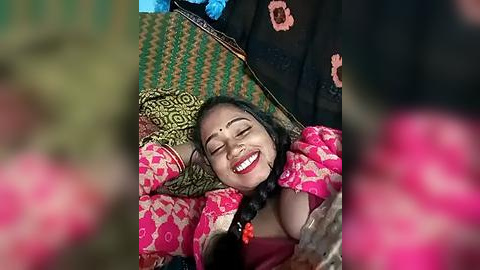 Media: Video of a smiling Indian woman in traditional pink and green saree, braided hair, and bindi, lying on colorful fabric, surrounded by blurred people.
