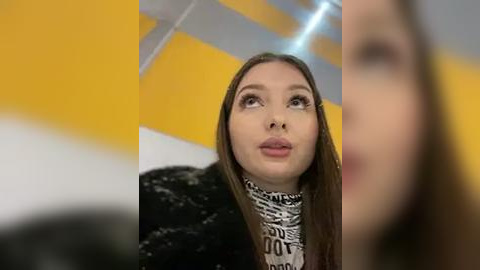 Media: Video of a young woman with long brown hair, wearing a black and white patterned scarf, looking up thoughtfully in a modern indoor setting with yellow and white geometric patterns on the walls.