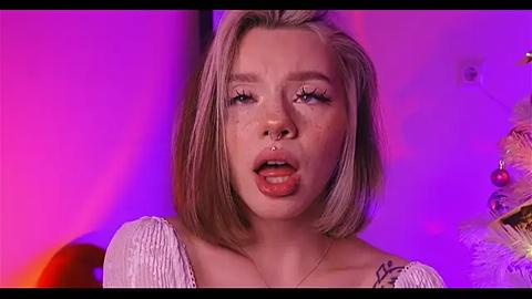 Media: Video of a young Caucasian woman with light skin and straight blonde hair, wearing a white, ribbed top. She has a nose ring and is licking her lips, set against a pink and purple backdrop.