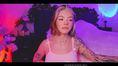 Media: A video of a young woman with blonde hair and tattoos, wearing a pink tank top, sitting in a bedroom with purple and red lighting, Christmas decorations, and a moon-shaped light fixture.