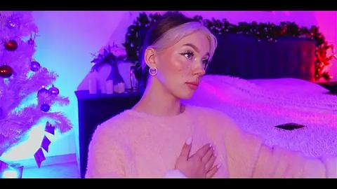 Media: Video of a young woman with fair skin and blonde hair, wearing a fluffy white sweater, standing in a cozy, dimly lit bedroom with Christmas decor.